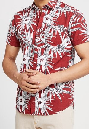 Homme portant une chemise rouge à manches courtes avec un imprimé floral tropical blanc, debout les mains jointes à la taille, pantalon beige visible sous la chemise.