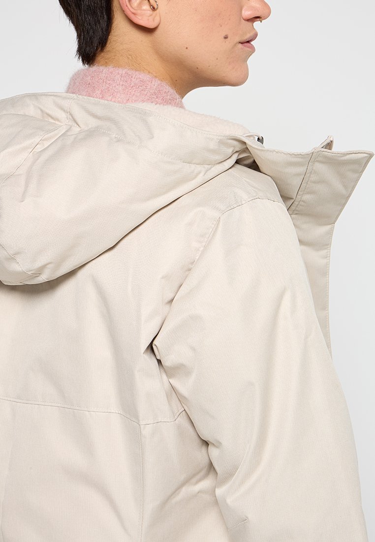 Beige wasserdichte Jacke mit Kapuze, die eine glatte Textur und ein elegantes Design aufweist. Sie hat einen hohen Kragen und einen Reißverschluss.