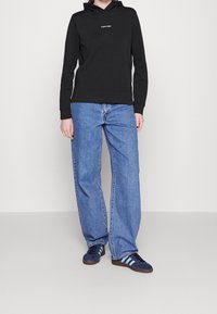 Felpa nera con logo frontale, abbinata a jeans larghi di un blu chiaro e sneakers blu scuro con tre strisce bianche.