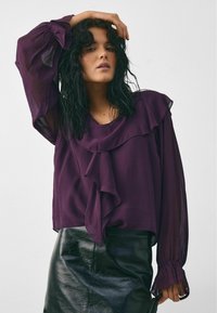 Kvinde med krøllet mørkt hår iført en gennemsigtig lilla bluse med flæser og en sort lædernederdel, der poserer med den ene hånd på sit hoved.