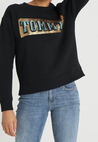 Czarna bluza z fakturą prążkowaną, z dużym logo "TOMMY" w kolorach morskim i złotym na przodzie. Casualowy design, luźny krój.