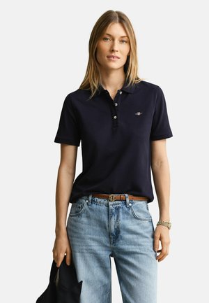 Femme portant un polo bleu marine foncé, un jean bleu clair avec une ceinture marron, tenant une veste sombre dans sa main gauche, sur un fond uni.