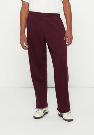 Hombre vestido con pantalones de chándal burdeos de Adidas y zapatillas blancas, de pie contra un fondo blanco liso.