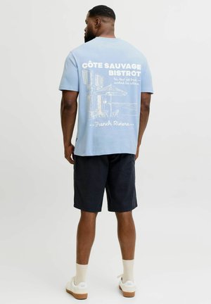 Bărbat purtând un tricou albastru deschis cu o ilustrație și text cu tema bistroului de pe Coasta de Azur, asortat cu pantaloni scurți negri, șosete albe și adidași albi.