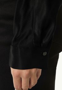 Black long-sleeve shirt with a smooth texture, featuring a folded cuff and a black button accent, closely cropped at the wrist.
