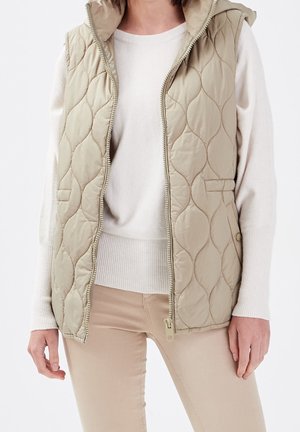 Veste matelassée beige sans manches avec capuche portée sur un pull blanc à manches longues et un pantalon beige ajusté, fermeture éclair partiellement ouverte.