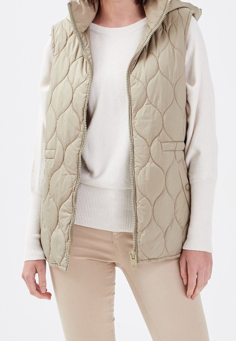 Veste matelassée beige sans manches avec capuche portée sur un pull blanc à manches longues et un pantalon beige ajusté, fermeture éclair partiellement ouverte.