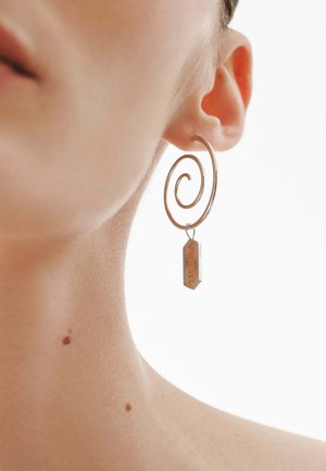Oreja de una persona con un pendiente de plata en forma de espiral, del que cuelga un pequeño colgante rectangular naranja, sobre un fondo blanco.