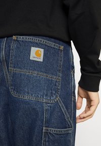 Pantalones cargo de mezclilla en azul oscuro con un acabado texturizado. Las características incluyen grandes bolsillos y un parche con el logo de Carhartt en naranja en la parte trasera.