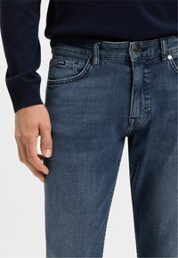 Dark blue denim jeans with a subtle texture, five-pocket design, and a branded tag. Close-up view shows the waistband and hand resting nearby.