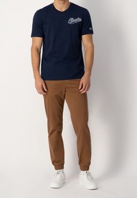 Muž v tmavě modrém tričku Champion, hnědých teplácích se elastickými manžetami a bílých teniskách, stojící na jednolitém pozadí.