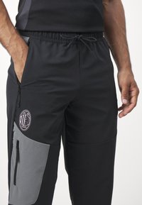 Zwarte sportbroek met een verstelbare tailleband, zakken aan de zijkanten en grijze accenten. Heeft een geborduurd logo op de linkerdij.