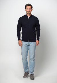 Cardigan zip-up in maglia di colore navy con colletto alto, maniche aderenti e polsini a costine, abbinato a jeans blu chiaro e sneakers grigie.
