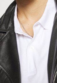 Osoba v bílém polokošili pod černou koženou bundou, viditelný stříbrný náhrdelník kolem krku.