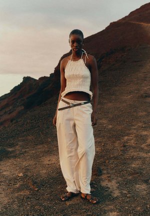 Femme debout sur un terrain rocailleux, portant un top blanc dos-nu, un pantalon crème avec une ceinture et des sandales cloutées, avec des collines et un ciel nuageux en arrière-plan.