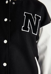 Veste varsity noire avec des accents blancs. Présente un "N" brodé texturé sur le devant, des boutons-pression et des manches grises contrastantes.