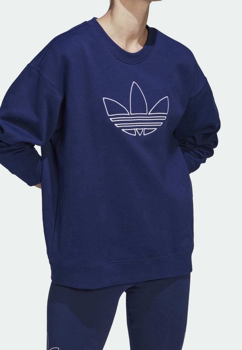 Osoba v volném modrém svetru s velkým bílým konturovaným logem Adidas trefoil na přední straně a odpovídajícími modrými kalhotami.