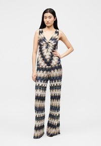 Γυναίκα που φοράει ολόσωμη φόρμα χωρίς μανίκια, με V-λαιμόκοψη και σχέδιο ζιγκ-ζαγκ σε μπλε, μπεζ και λευκό, στέκεται με το ένα χέρι στη μέση της.