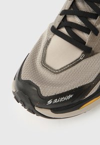 Αθλητικό παπούτσι από διχτυωτό υλικό σε μπεζ με μαύρες λεπτομέρειες. Διαθέτει υφή στην επιφάνεια, ενισχυμένη μύτη και ορατό σύστημα κορδονιών.