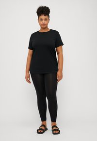 Black short-sleeve t-shirt, paired with black leggings. Model stands in black sandals. Smooth fabric and relaxed fit characterize the clothing.