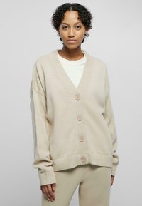 Beige knitted cardigan with a V-neck, five buttons down the front, ribbed cuffs and hem, and a relaxed fit. Plain background.