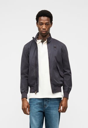 Νεαρός άνδρας που φοράει σκοτεινό γκρι μπουφάν με φερμουάρ πάνω από λευκό πόλο μπλουζάκι και μπλε τζιν, στέκεται μπροστά σε απλό φωτεινό φόντο.