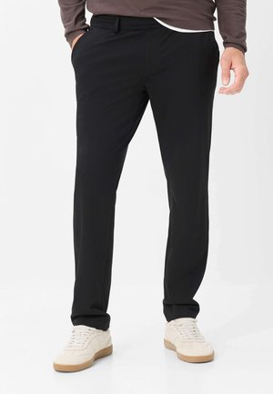 Mand iført sorte bukser med lige ben, brun sweater og hvide sneakers står med den ene hånd i lommen mod hvid baggrund.