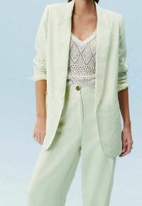 Femme portant un blazer jaune pâle et un pantalon à jambes larges avec un haut en maille blanche à dentelle, debout sur un fond bleu clair.