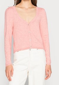 Rosa stickad kofta med V-ringning, långa ärmar och knäppt front. Har en mjuk textur och ribbad nederkant. Bärs över vita byxor.