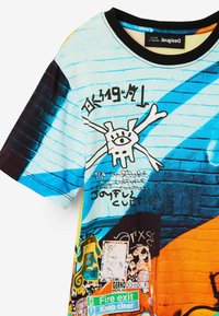 Kortærmet t-shirt i bomuldsblanding med livlige blå, sorte og orange farver. Indeholder grafitti-inspirerede grafikker og en tekstureret murstensmønster.