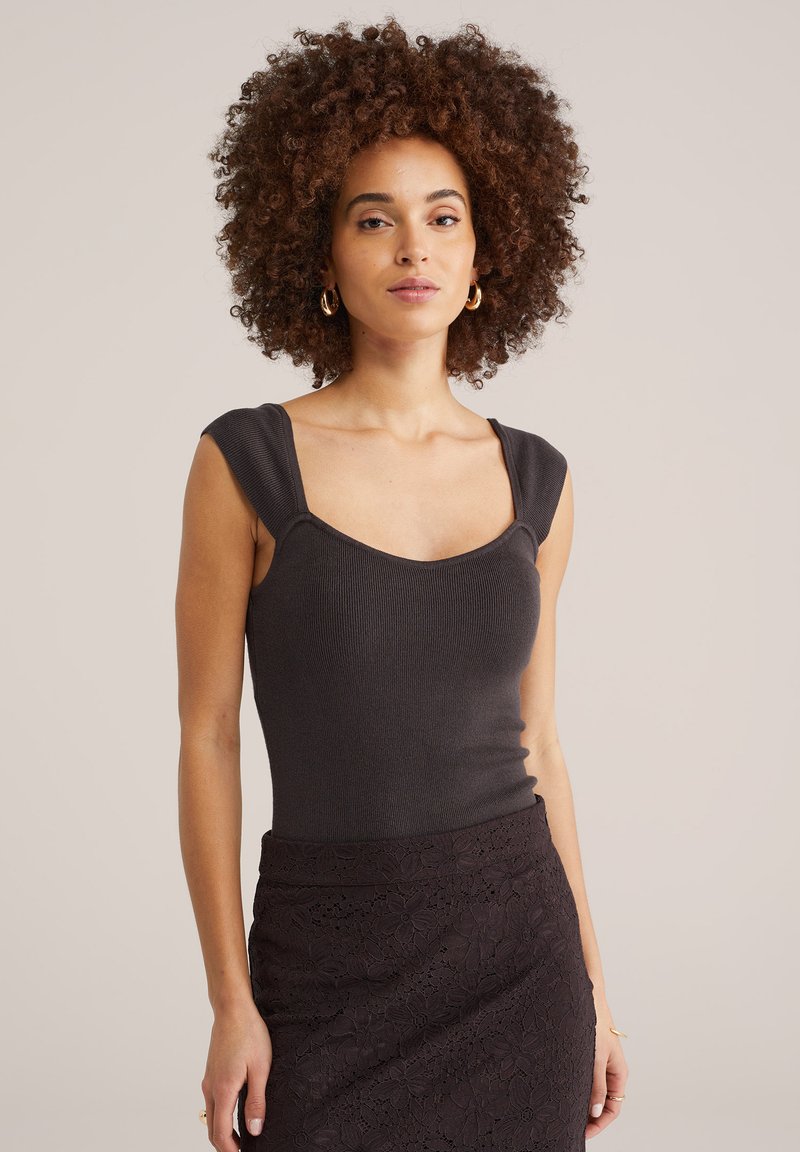 Femme aux cheveux bouclés portant des boucles d'oreilles créoles dorées, un haut noir sans manches côtelé et une jupe en dentelle florale noire, debout devant un fond beige.