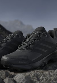 Paire de chaussures de randonnée noires imperméables avec tissu Gore-Tex et semelles robustes placées sur un terrain rocheux avec un arrière-plan montagneux flou.