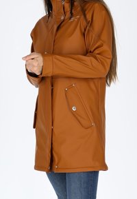 Femme portant une longue veste marron avec des poches à boutons-pression et un jean bleu, mains jointes devant, sur un fond blanc uni.