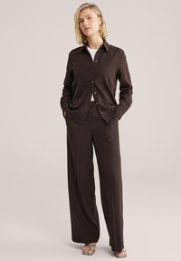 Vrouw met kort blond haar die een donkerbruine blouse met knopen en bijpassende wijde broek draagt, staand met handen in de zakken.