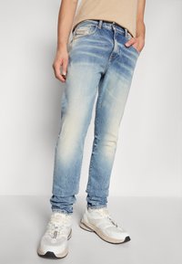 Vaaleansiniset farkut, joissa on haalistettu pesu, kapea leikkaus, viisi taskua ja hienovaraiset ompeluhelmet, yhdistetty valkoisiin tennareihin.