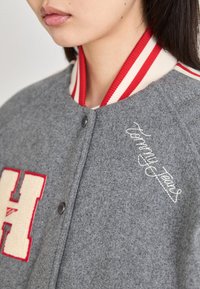 Pilkas vilnonis švarkas su raudonais ir baltais dryžuotais ribiniais apykaklėmis, turi didelį raštą "H" ir išsiuvinėtą "Tommy Jeans" logotipą.