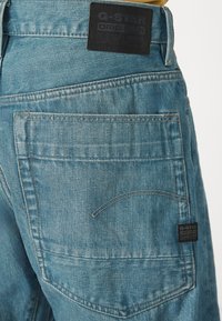 Vaqueros de mezclilla en azul claro con un acabado texturizado, que presentan un diseño de costura en el bolsillo trasero y una etiqueta negra de G-Star en la cintura.