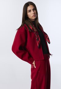 Chaqueta roja corta con mangas largas, que cuenta con una bufanda adjunta con un patrón en negro y rojo. Combinada con pantalones de cintura alta rojos a juego.