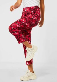 Person som bär röda mönstrade byxor och vita klumpiga sneakers, lyfter ena benet mot en enkel ljus bakgrund.