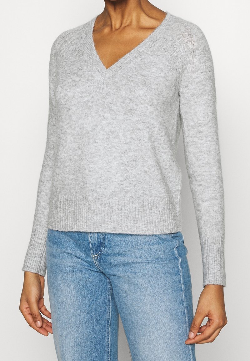 Femme portant un pull à manches longues gris clair à col en V et un jean bleu, debout avec les mains détendues le long du corps.