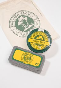Lata de cera de zapato verde con etiqueta amarilla, bloque de cuidado gris con detalles amarillos, y una bolsa de algodón beige con un logo impreso.