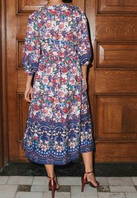 Maxikleid mit Blumenmuster und Puffärmeln, in Marineblau mit bunten Blumen und einem gestuften Saum, kombiniert mit metallischen Absatzsandalen.