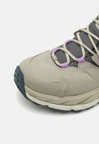 Vaaleanbeige vaelluskenkä, jossa on teksturoitu pinta, violetit nauhaaksentit ja kova musta kumiulkopohja pitoa varten. Verkko-osat hengittävyyttä varten.