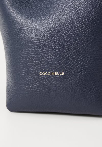 Pochette en cuir bleu marine avec une surface texturée. Présente un subtil logo doré "Coccinelle" à l'avant. Design simple et compact.
