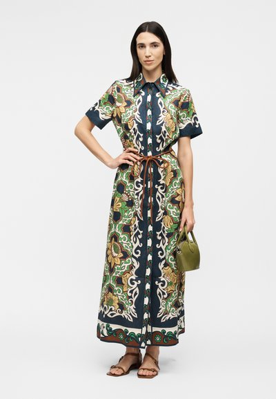 Γυναίκα με μακρύ, εμπριμέ φόρεμα σε πράσινες, ναυτικές και μπεζ αποχρώσεις με floral σχέδια, φορώντας καφέ σανδάλια και κρατώντας μια μικρή τσάντα ελιάς σε σκούρο πράσινο χρώμα.