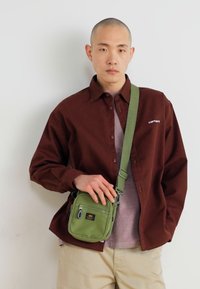 Camicia marrone con bottoni, top grigio, pantaloni khaki e borsa a tracolla verde con zip ed emblema. Tessuto liscio e forma rettangolare della borsa.