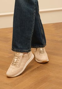 Sneaker in suede beige con suola bianca, dotati di chiusura con lacci e zip, abbinate a jeans a gamba larga blu scuro su un pavimento in legno.