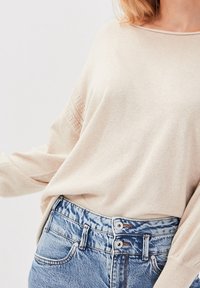 Nainen, joka on pukeutunut vaaleanbeigeen pitkähihaiseen neulepaitaan, joka on tuotu ylös korkea vyötäröisten sinisten farkkujen sisään, joissa on kaksoispainike.