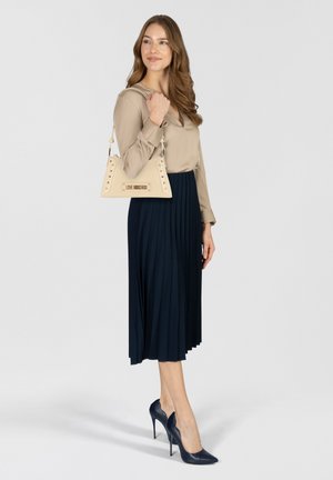 Femme portant un chemisier beige, une jupe midi plissée bleu marine, des escarpins pointus bleu marine et portant un sac à main beige à clous sur l'épaule.