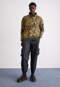 Fleece-pullover med camo-mønster i brunt og grønt, med glidelås-kollaring; kombinert med løstsittende svarte cargo-bukser og kraftige svarte støvler.